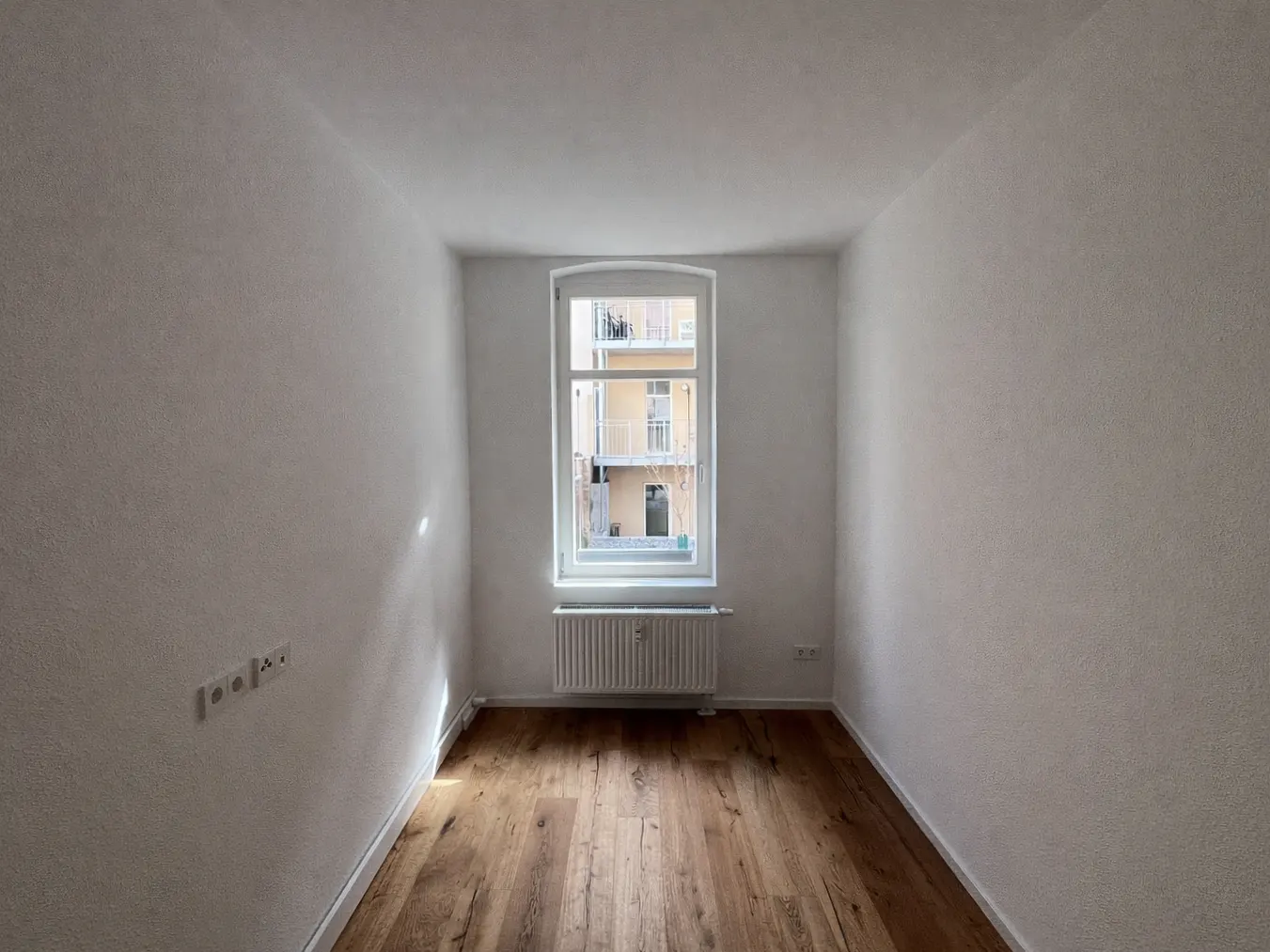 Empty room with hardwood floors, white walls, and a window. A radiator sits beneath the window, which looks out onto a building.