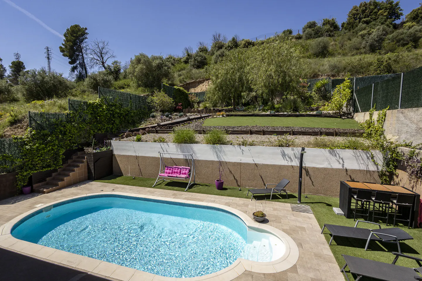 A backyard with a blue pool, lounge chairs, a swing with pink cushions, and a bar table, set against a green hillside.