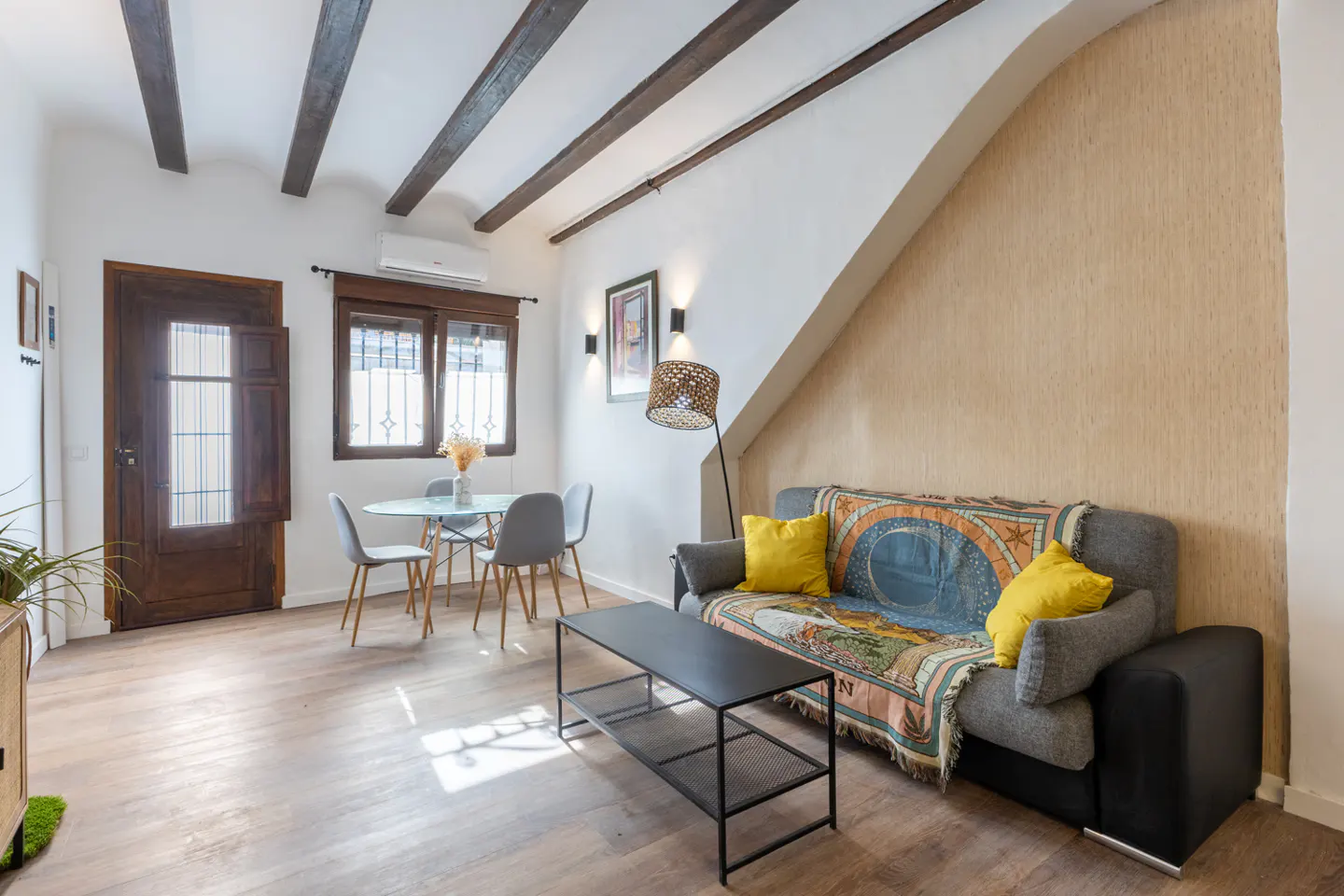 Living room with wood floors, exposed beams, and a gray sofa with yellow pillows. A round glass table and gray chairs are near a window.