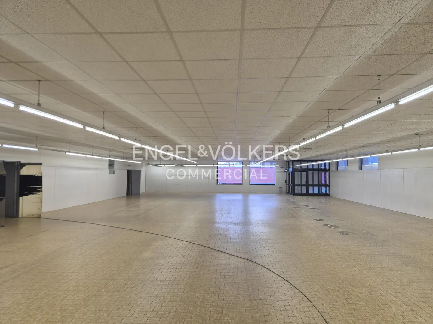 Wide, empty commercial space with white walls, tile floor, and fluorescent lighting. Engel & Völkers Commercial logo is superimposed in the center.