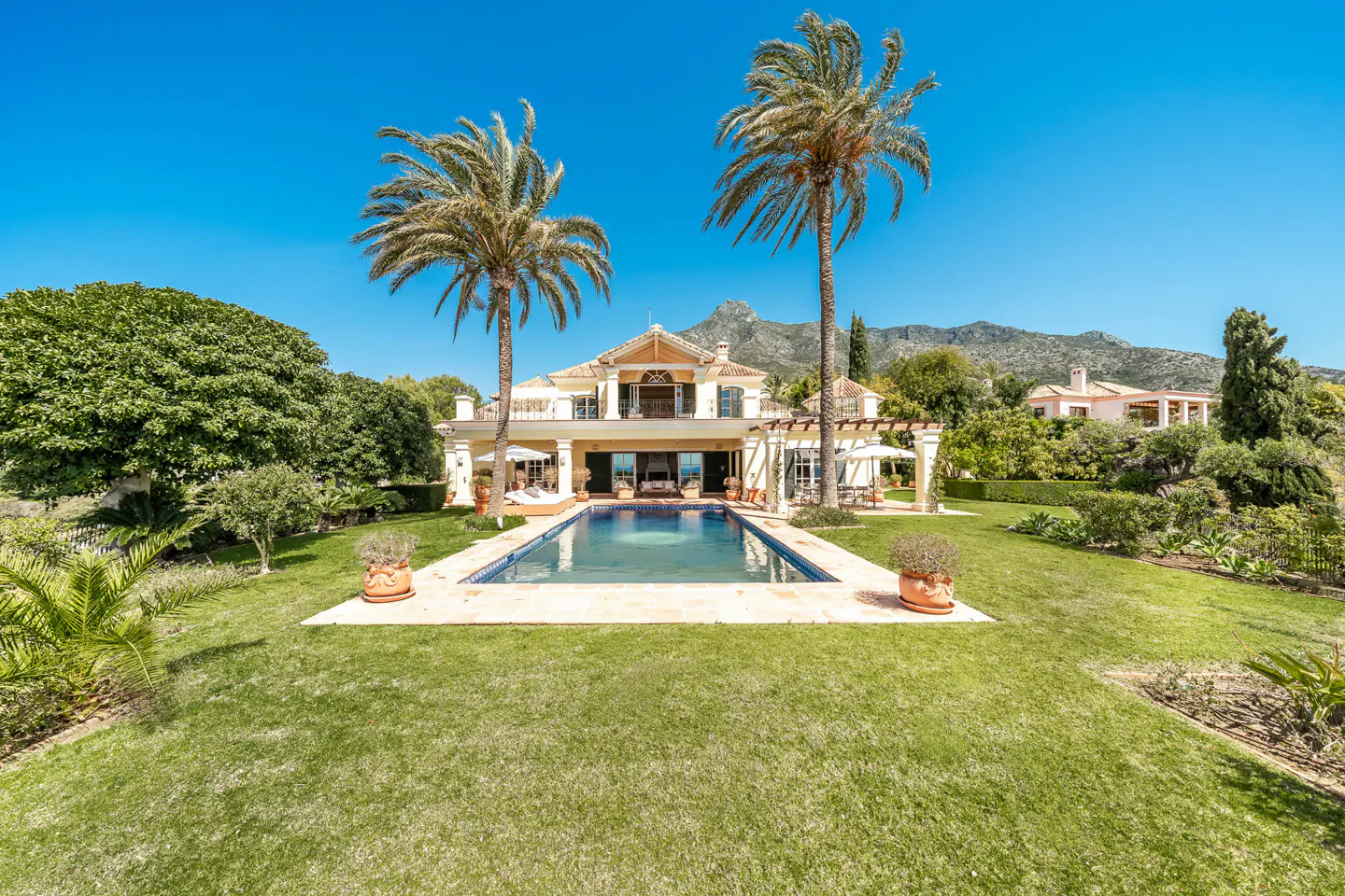 Luxury home with a pool, palm trees, and a manicured lawn under a clear blue sky. Mountains are visible in the background.