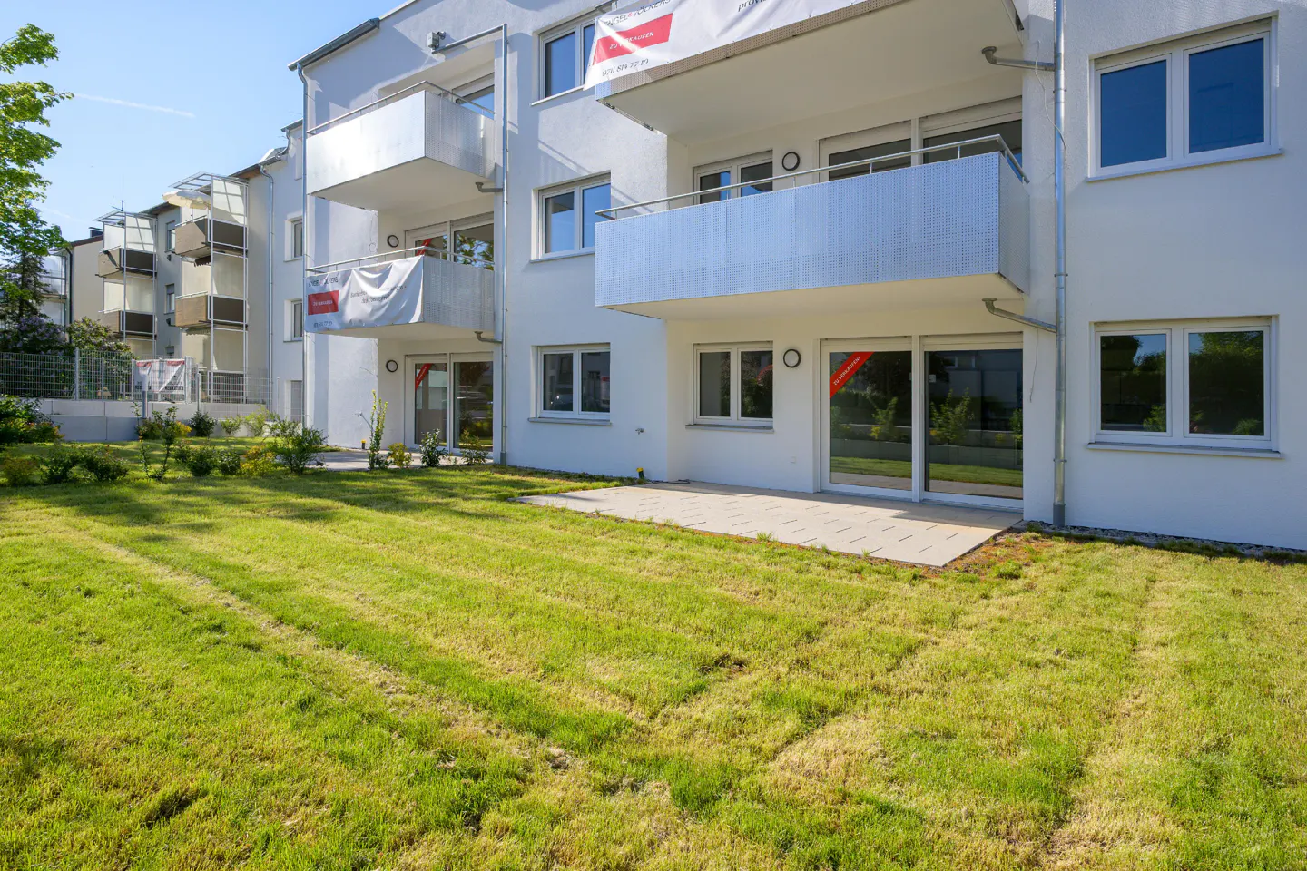 Modern white apartment building with balconies and a green lawn in front. Banners hang from some balconies.