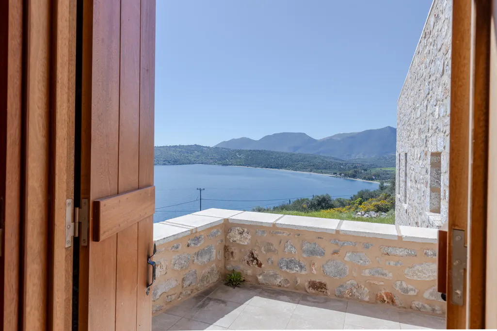 View from open wooden doors onto a stone balcony overlooking a blue bay with mountains in the distance under a clear blue sky.