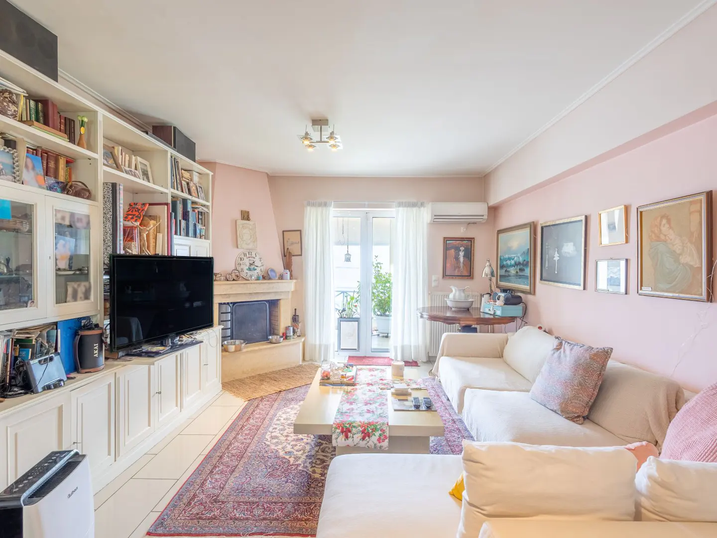 Bright living room with pink walls, white sofas, and a large floral rug. A fireplace and built-in bookshelves add character. Balcony doors let in natural light.
