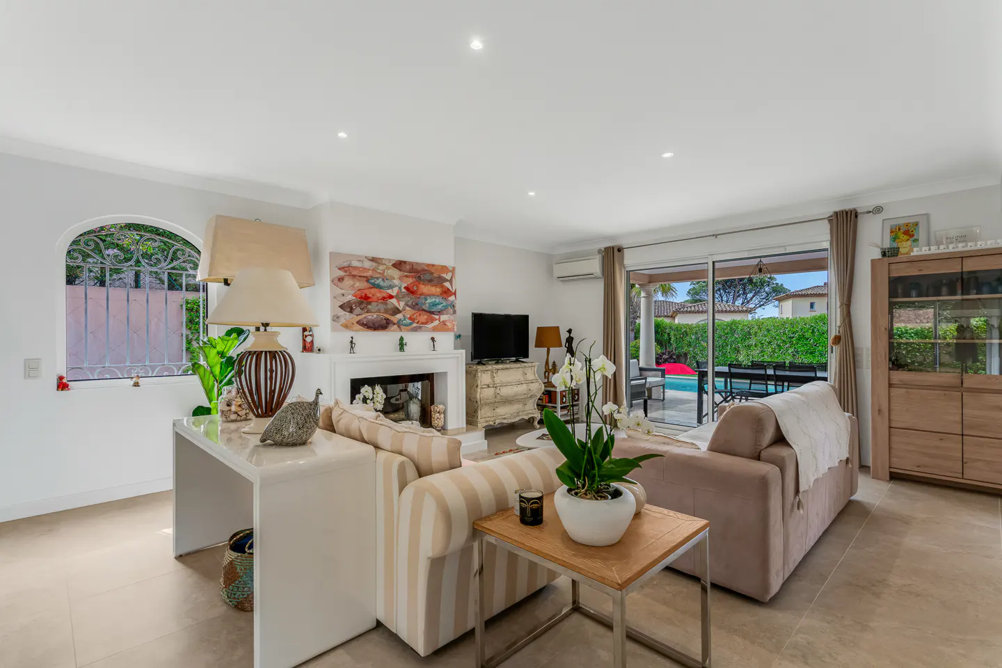 Bright living room with beige tile floor, striped sofa, and sliding glass doors to a pool area. A white fireplace and fish art add charm.