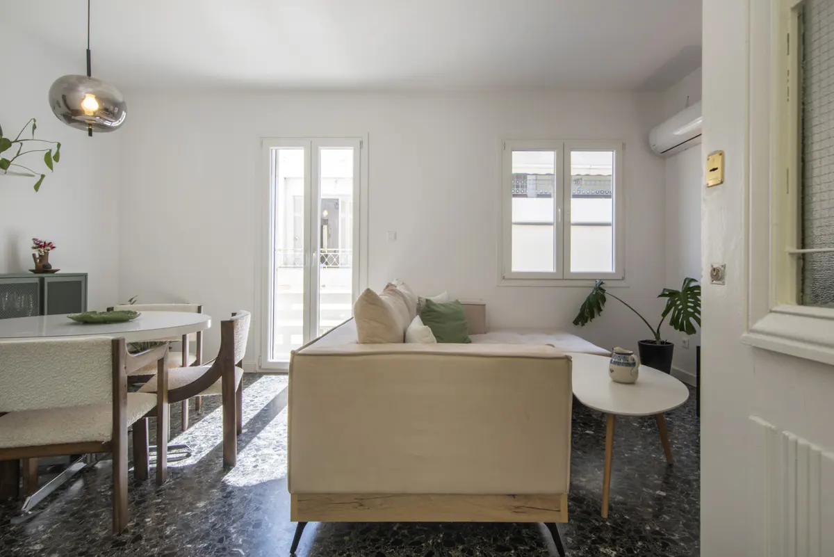 Bright living room with white walls, a beige sofa, round table, and black terrazzo floor. A glass pendant light hangs above the dining area.