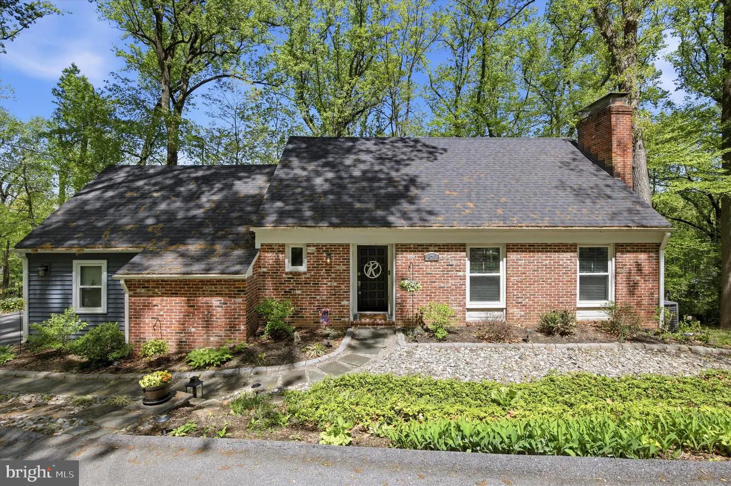 A single-story brick house with a dark roof and a brick chimney surrounded by green trees.
