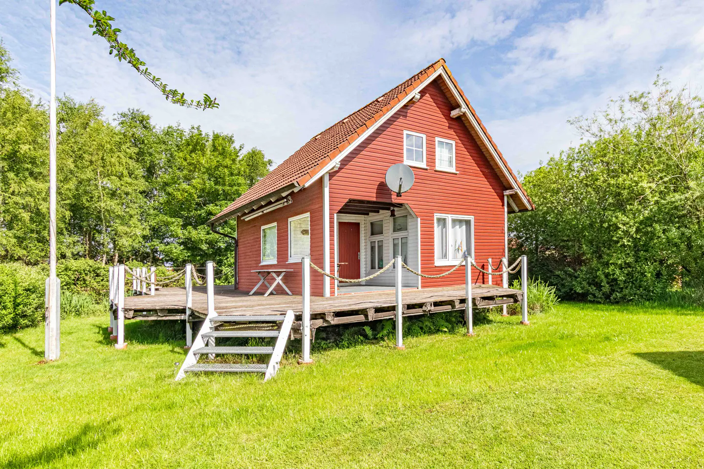 Red cottage-style house with a wooden deck, white railings, and green lawn. Trees and blue sky in the background.
