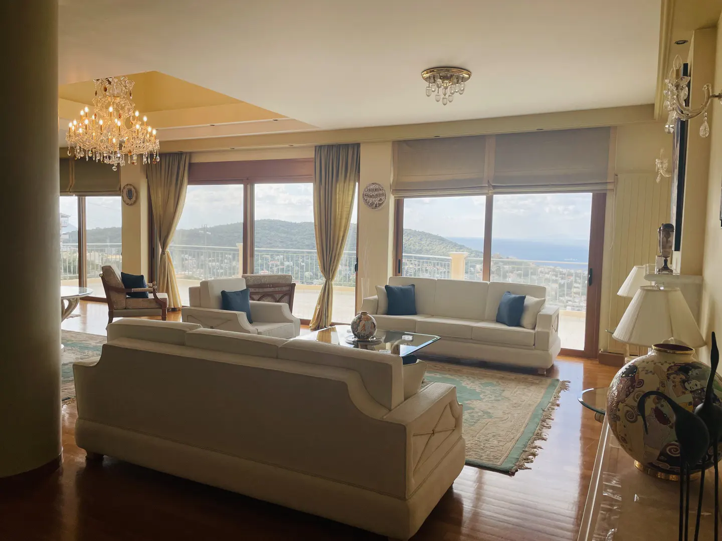 Bright living room with white sofas, blue pillows, and a view of the city and sea through large windows. A crystal chandelier hangs from the ceiling.