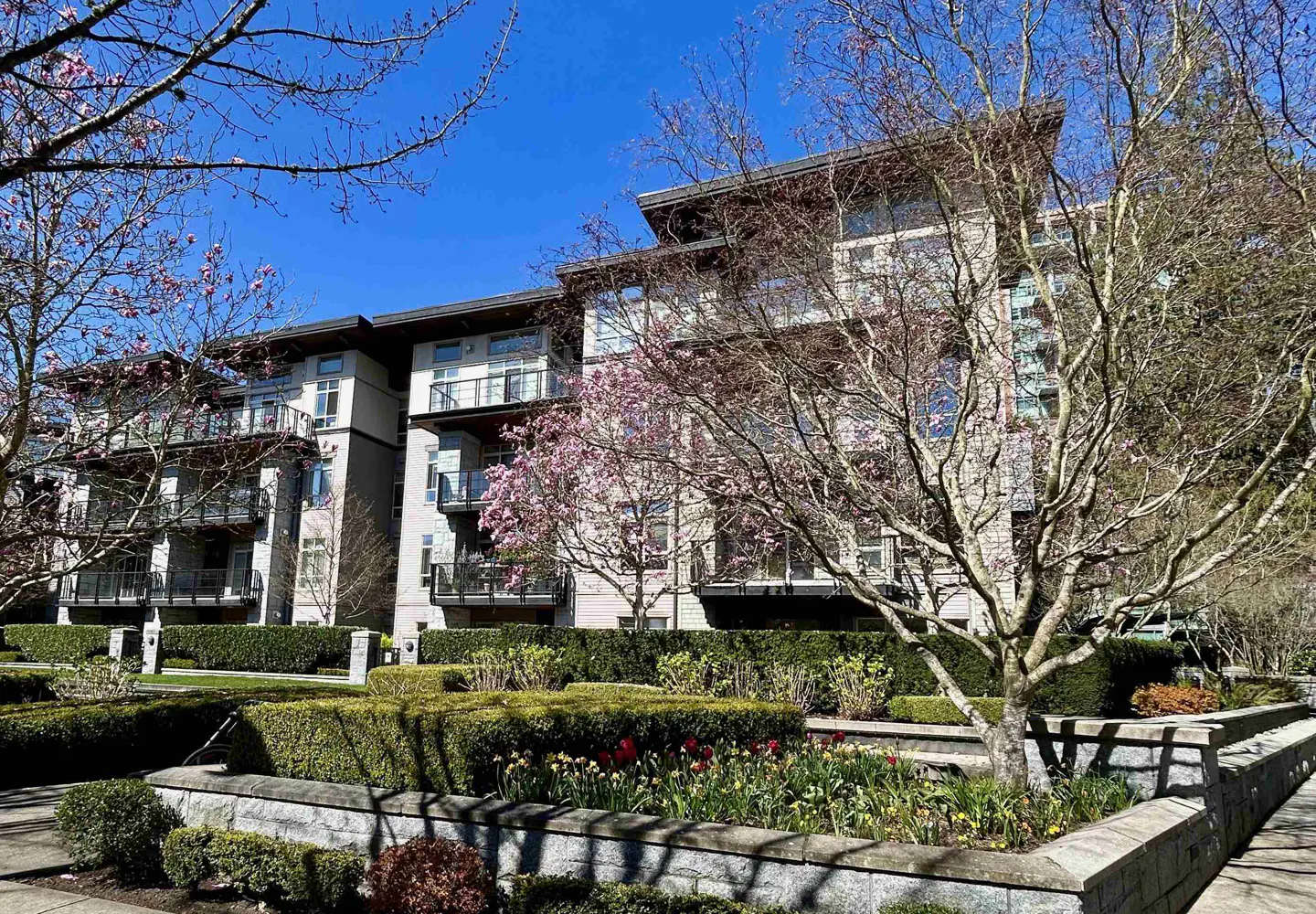 Three-story condo building with balconies, surrounded by manicured hedges, trees, and a flower bed under a clear blue sky.