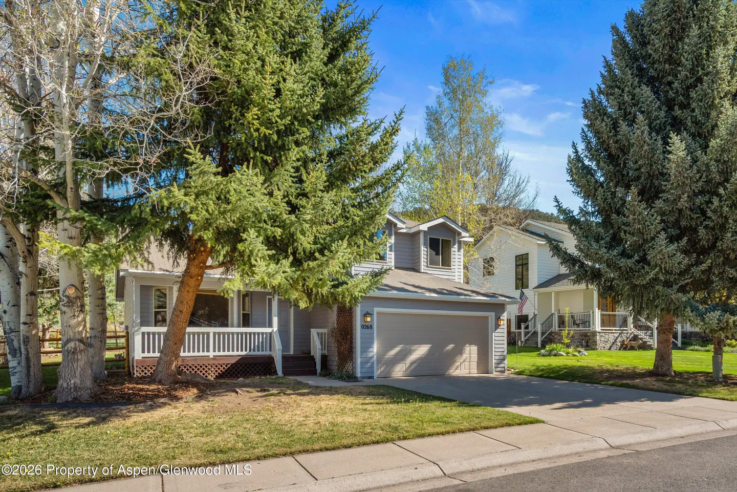 A light blue house with a front porch and an attached garage, surrounded by green trees.