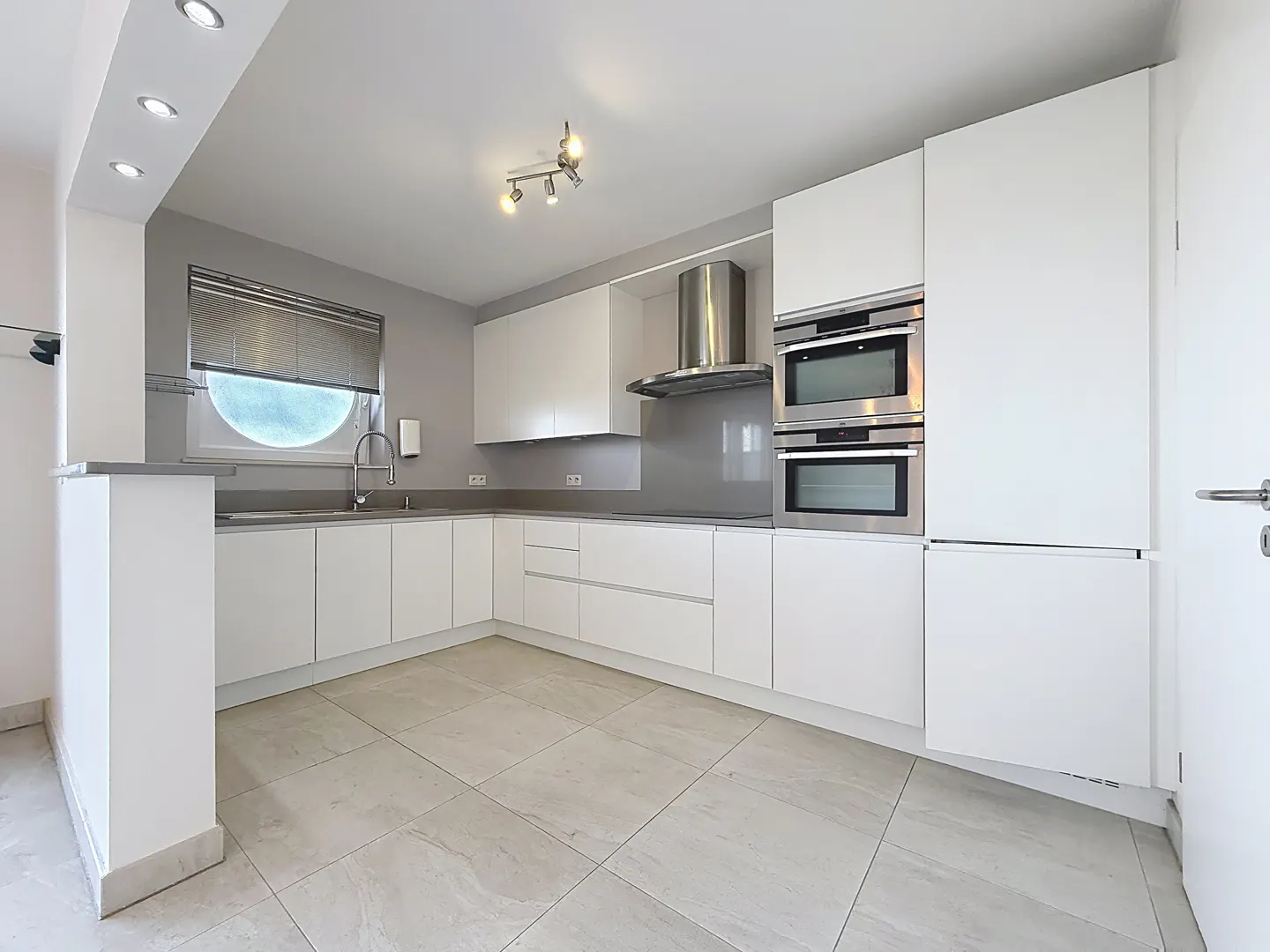 Bright, modern kitchen with white cabinets, stainless steel appliances, and light tile flooring. A round window adds unique detail.