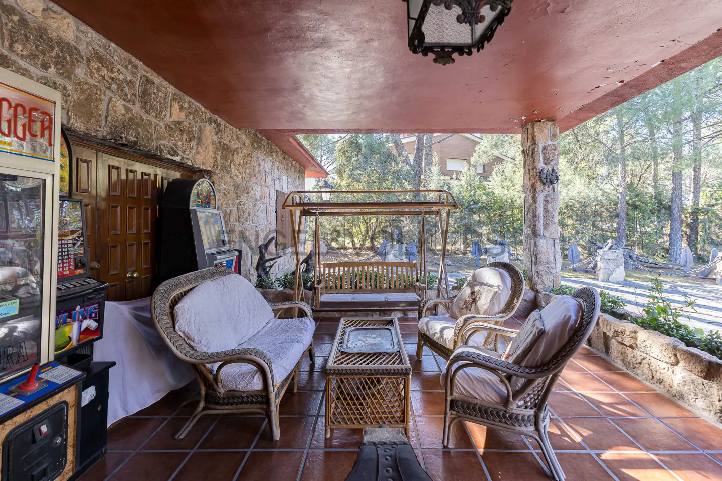 Covered patio with stone walls, terracotta tile floor, and wicker furniture. A wooden swing hangs in the background, with arcade games visible on the left.