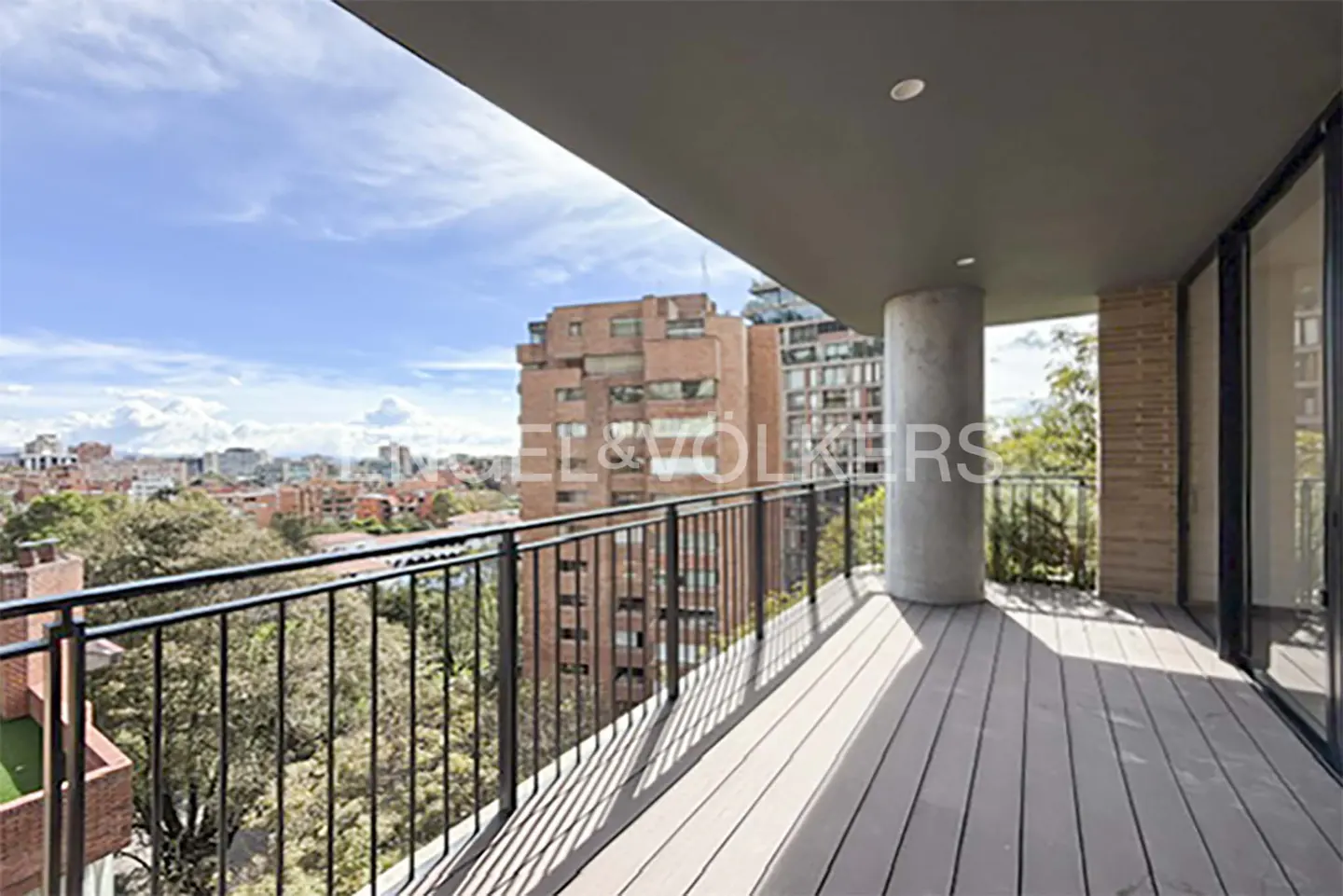 Balcony view with black railings, gray wood floor, and city skyline. A concrete pillar supports the gray ceiling. Trees and brick buildings are visible.