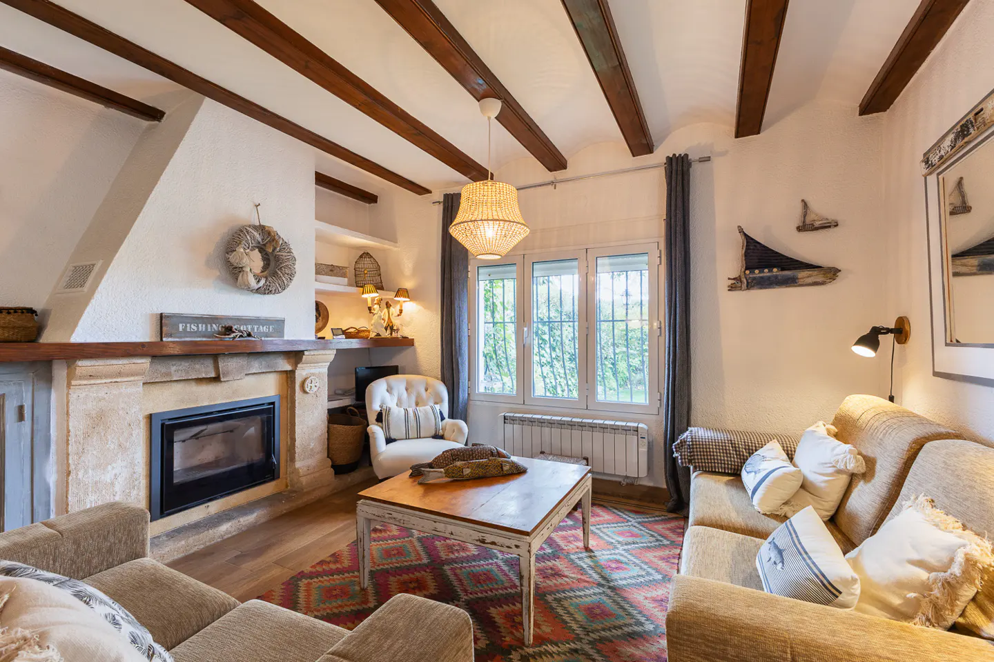 Cozy living room with stone fireplace, wood beams, and patterned rug. Neutral sofas and a white armchair create a comfortable space.