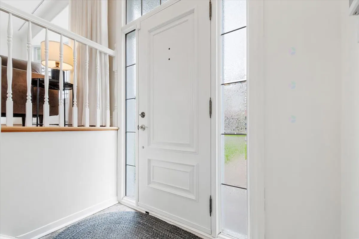 Bright entryway with a white front door flanked by sidelight windows. A gray rug sits at the entrance. A staircase with white railings is visible.