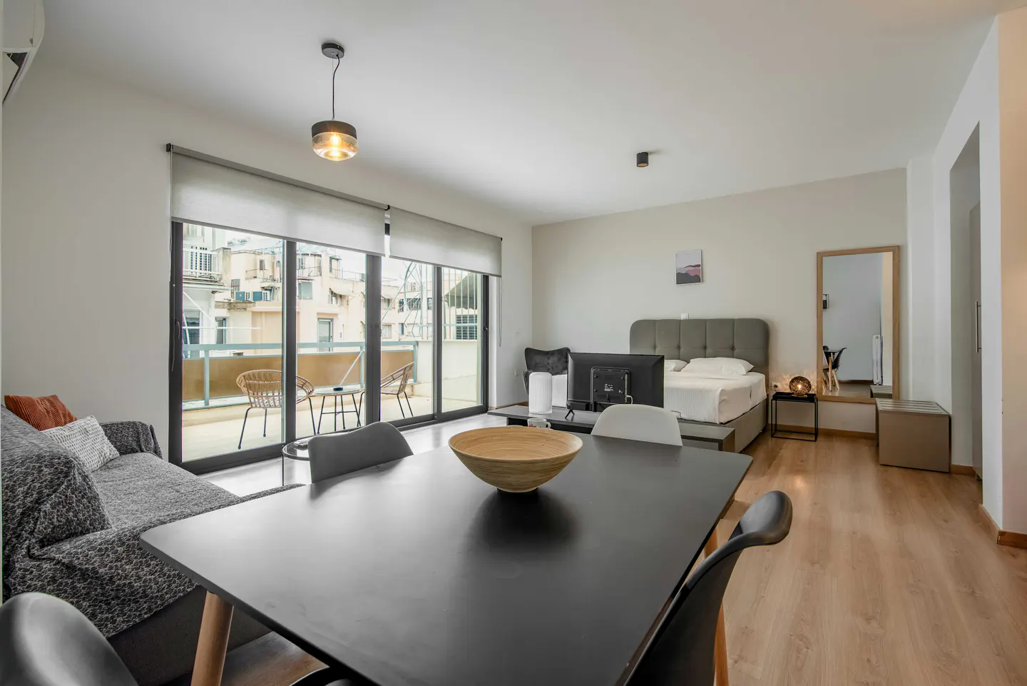 Bright studio apartment with a black dining table, gray sofa, and bed. Balcony with chairs and city view.