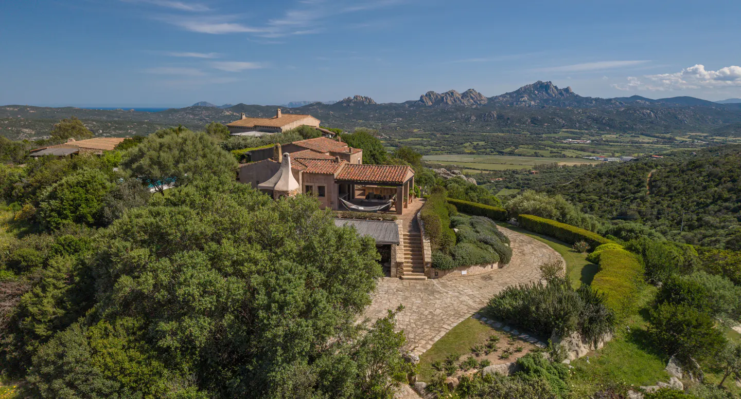 Aerial view of a terracotta-roofed house with a hammock on a hill, surrounded by green trees and a stone path, with mountains in the background.
