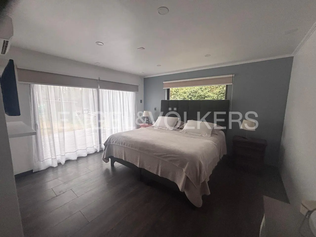 Modern bedroom with a king-sized bed, dark grey accent wall, wood-look tile floors, and large windows with sheer curtains.