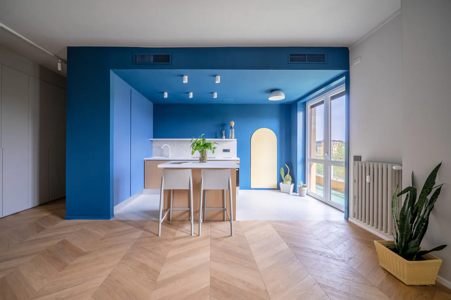 Bright apartment interior with blue walls, herringbone wood floors, and a kitchen island with two chairs. A yellow archway and large windows add to the modern design.