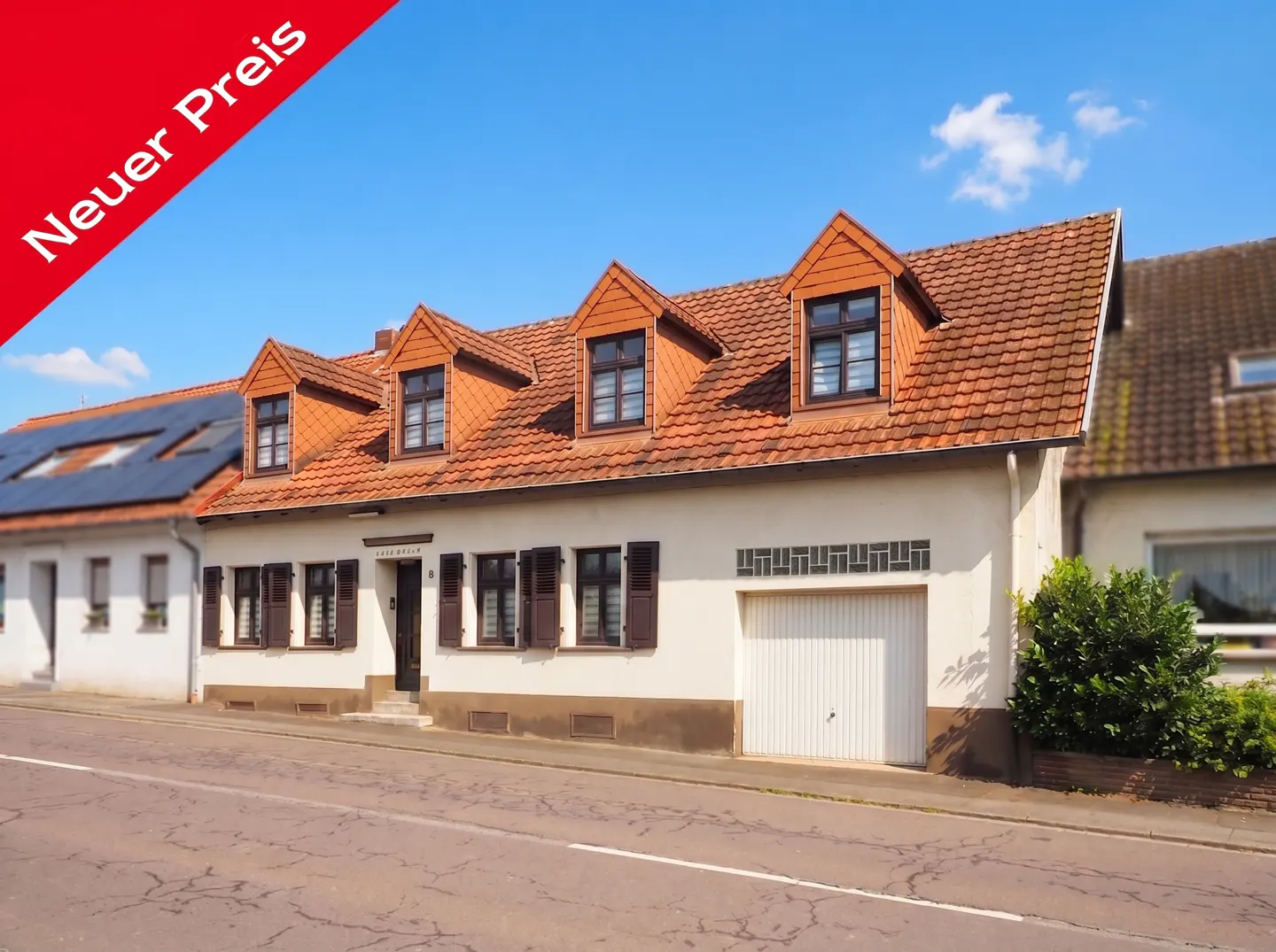 Exterior of a two-story white house with a red tile roof and brown shutters under a blue sky. A red banner reads "Neuer Preis."