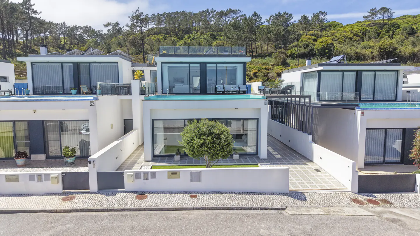 Modern white houses with pools on the roof, set against a green, tree-covered hillside under a blue sky.
