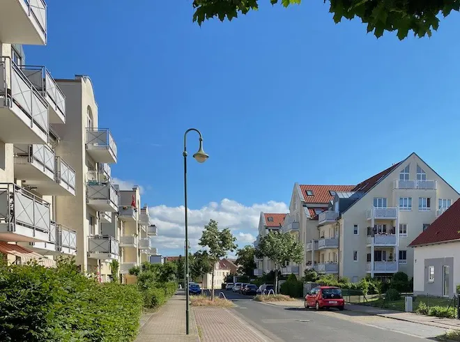 Street view of modern apartment buildings with balconies under a blue sky. A sidewalk and street lamp are in the foreground.