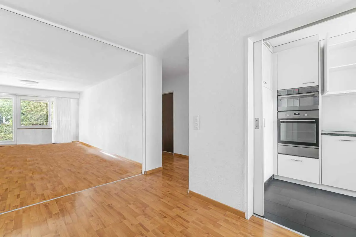 Bright, modern apartment interior with hardwood floors. White walls, large windows, and a glimpse of a white kitchen with stainless steel appliances.