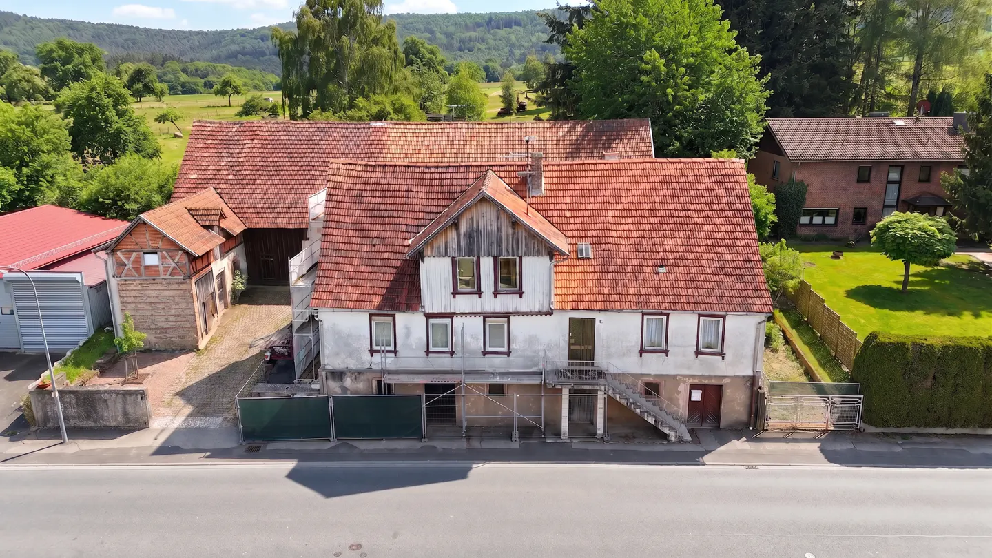 Two-story white house with a red tile roof and scaffolding in front, with a barn and green trees in the background.
