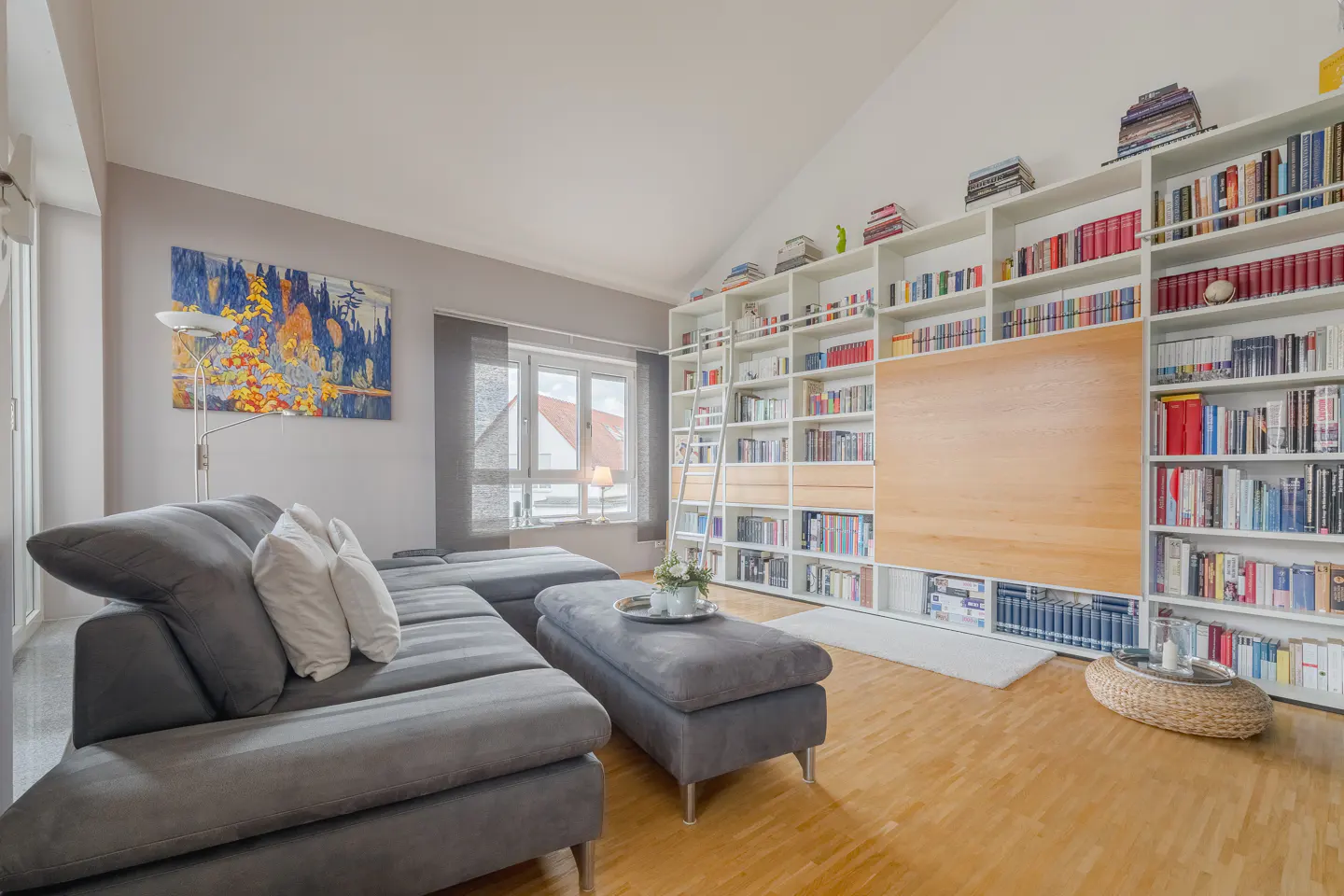 Bright living room with a gray sectional sofa, ottoman, and floor lamp. A large white bookshelf with a ladder fills the back wall.