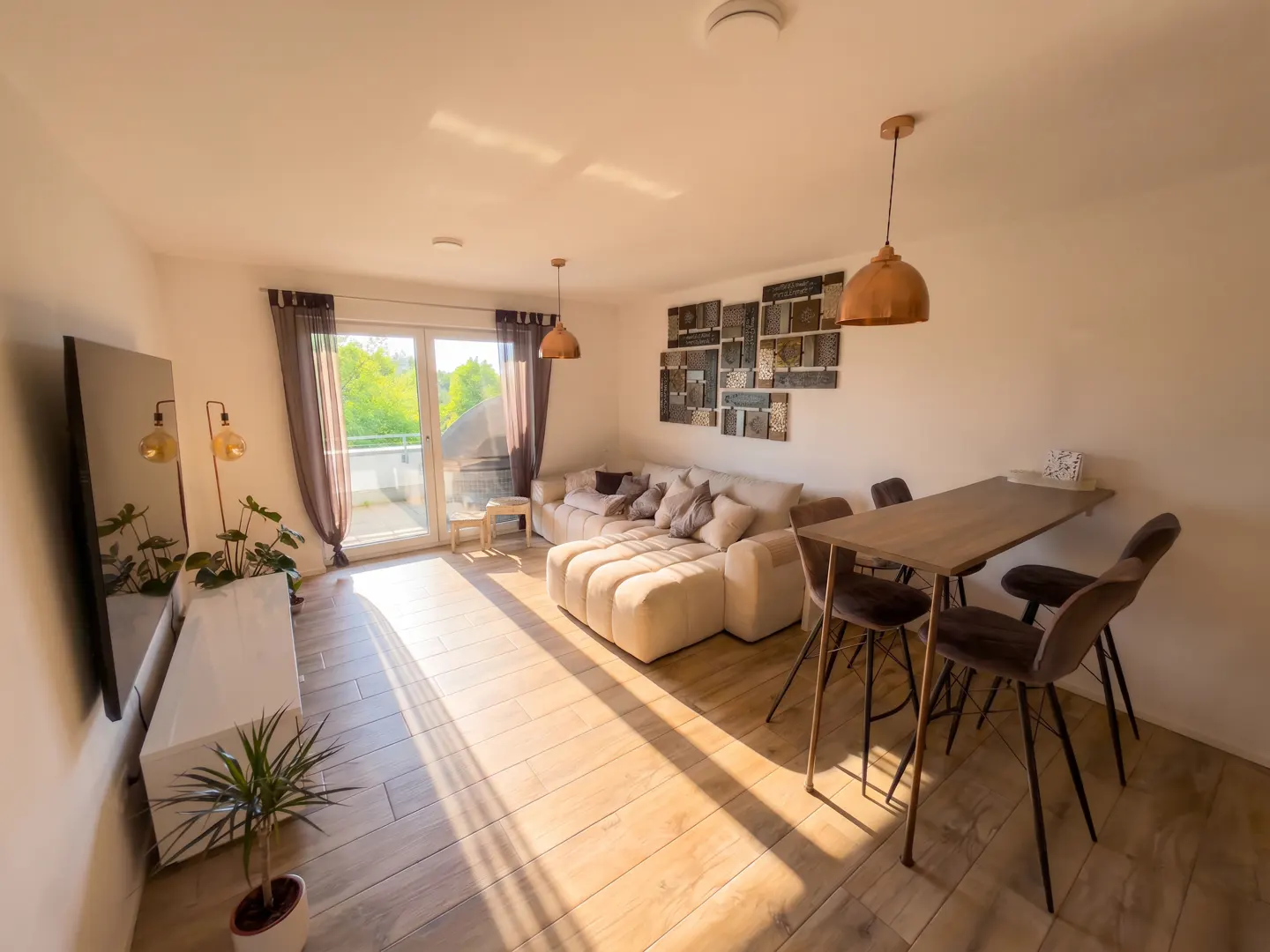 Sunlit living room with a beige sectional sofa, dining table with four chairs, and a balcony with a view of trees.