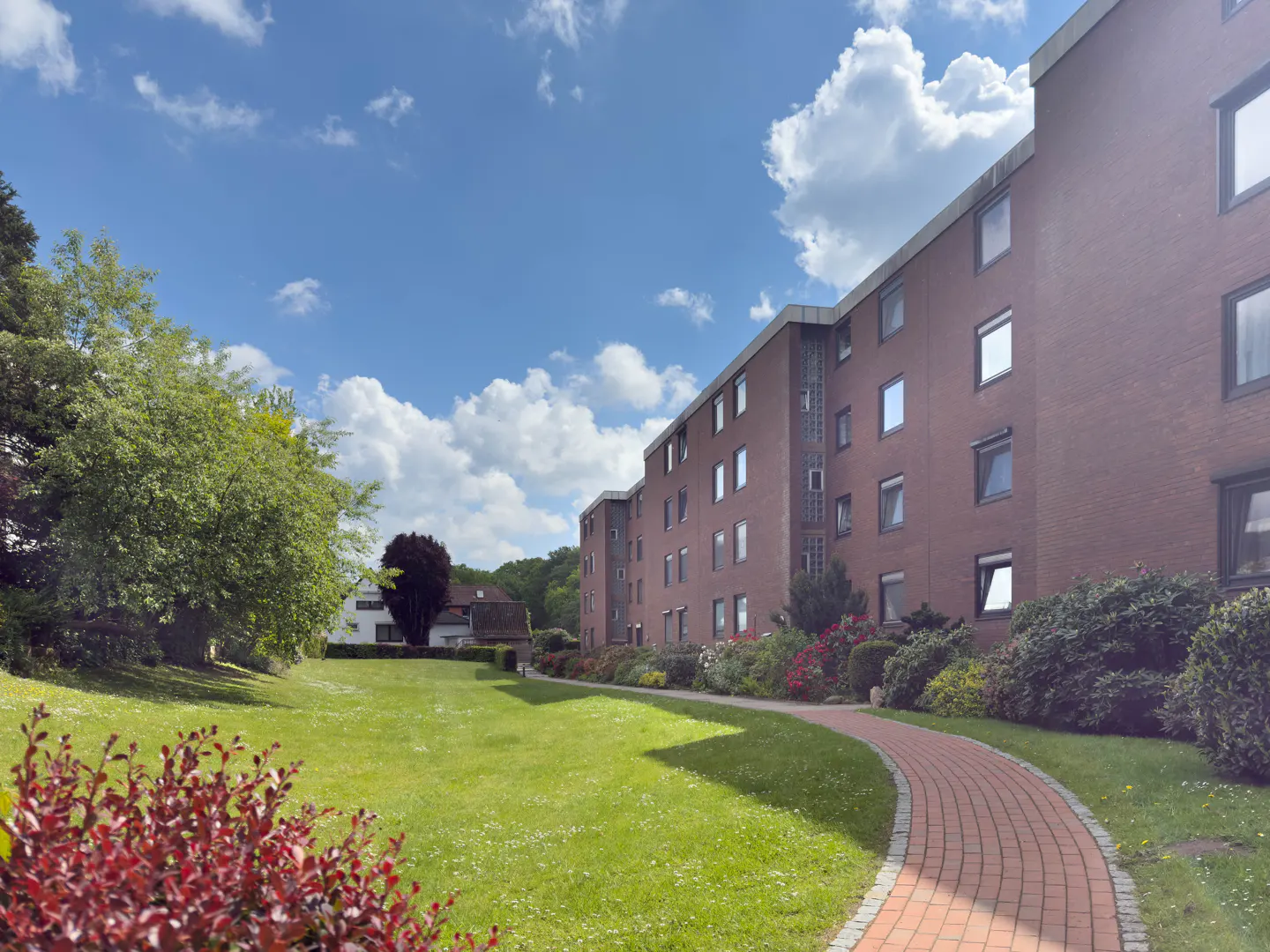 Brick apartment building with a curved brick walkway, green lawn, trees, and blue sky with clouds.