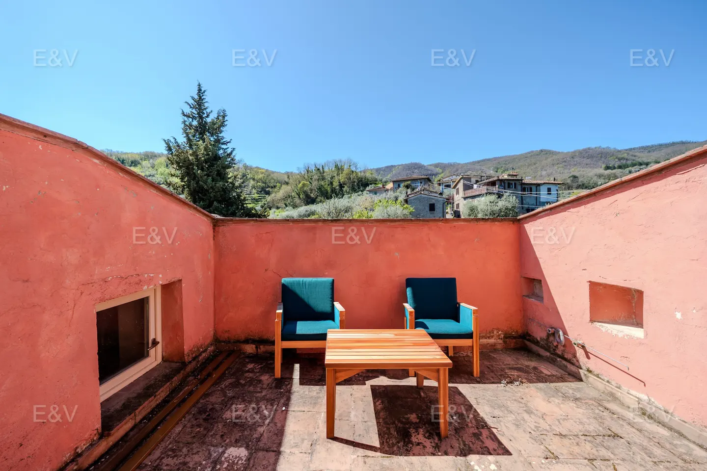 Outdoor patio with two blue cushioned chairs and a wooden table, surrounded by pink walls and a view of a hillside village.