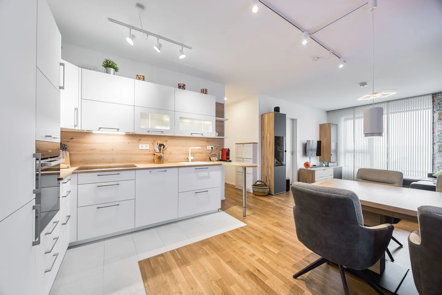 Bright, modern kitchen and dining area with white cabinets, wood accents, and hardwood floors. Gray chairs surround a wooden table.