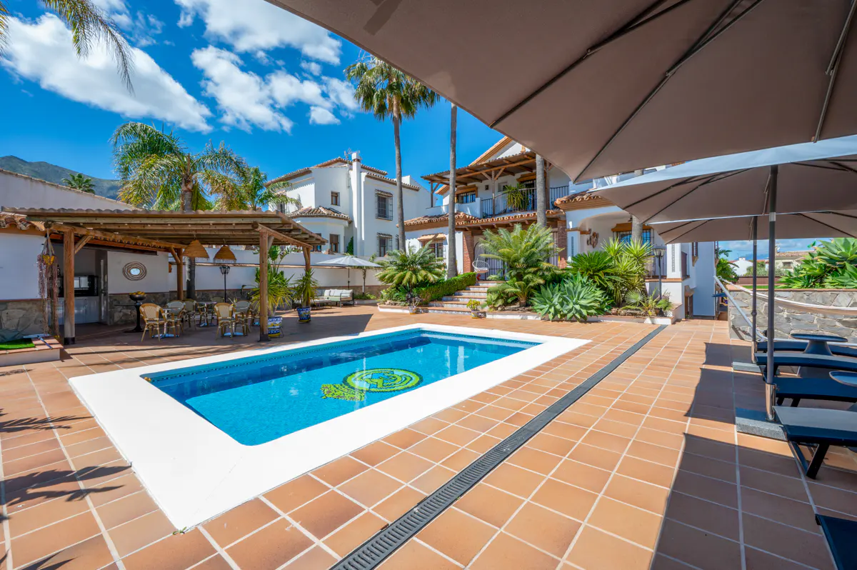 Outdoor pool area with blue water, surrounded by terracotta tiles, white buildings, palm trees, and patio umbrellas.