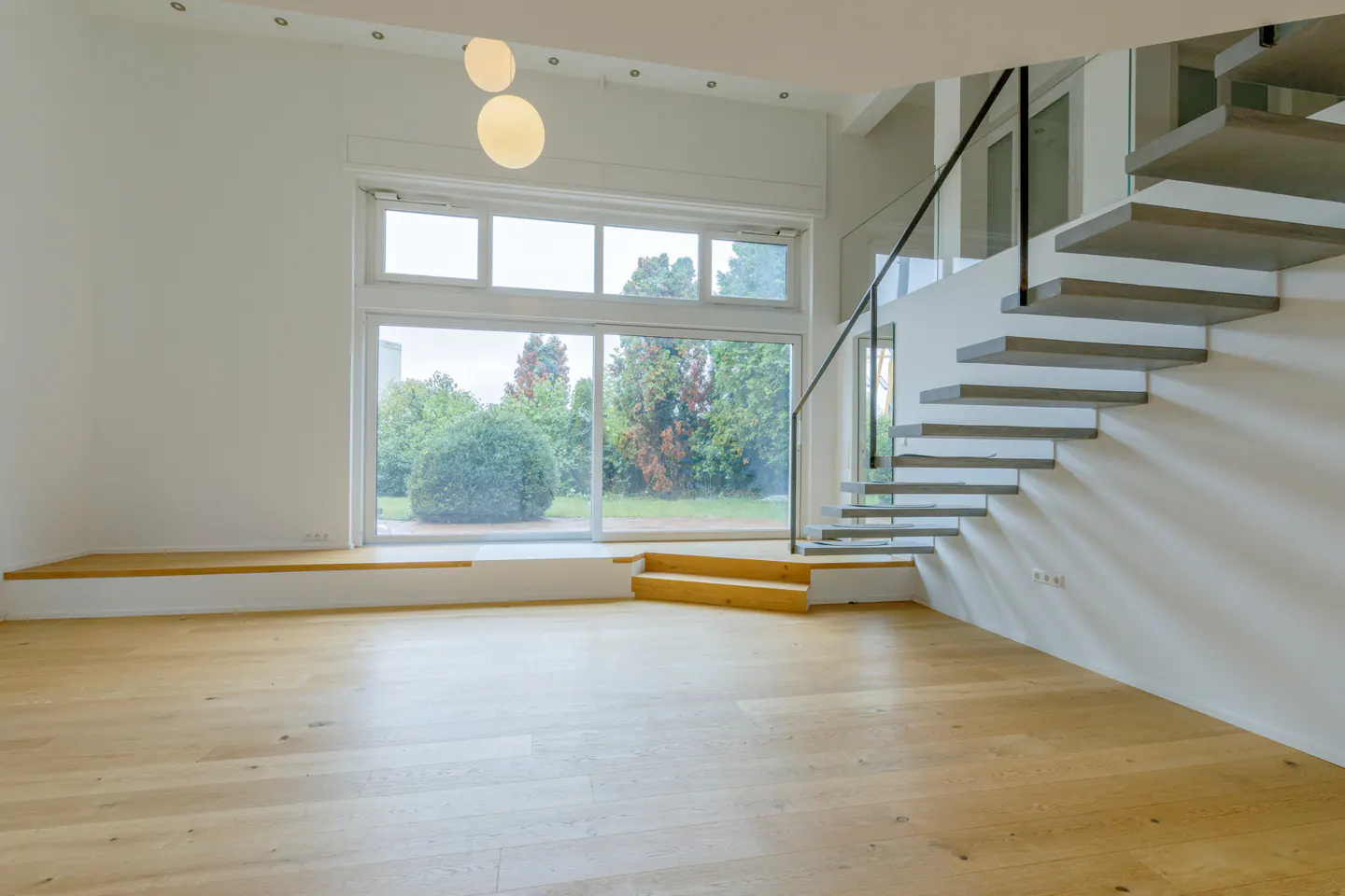 Bright, modern room with wood floors, white walls, and a large window overlooking a green garden. Floating stairs with black railing on the right.