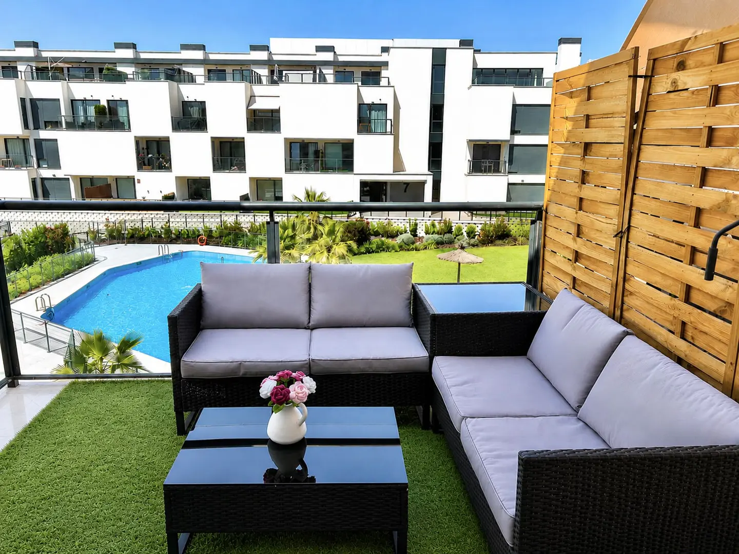 Modern balcony with wicker sofa, glass coffee table, and flower vase, overlooking a swimming pool and apartment complex.