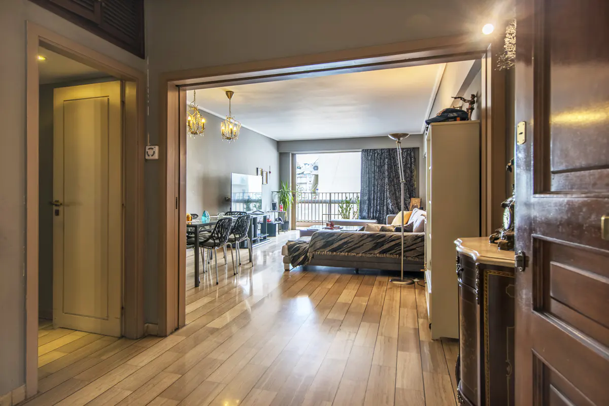 View from a hallway into a living room with hardwood floors, a dining set, a grey sofa, and a balcony with sheer curtains.