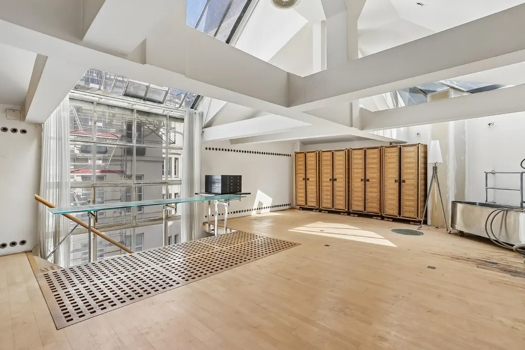 Bright, modern loft with skylights, white walls, and light wood floors. Wicker cabinets line the back wall. A glass table overlooks a large window.