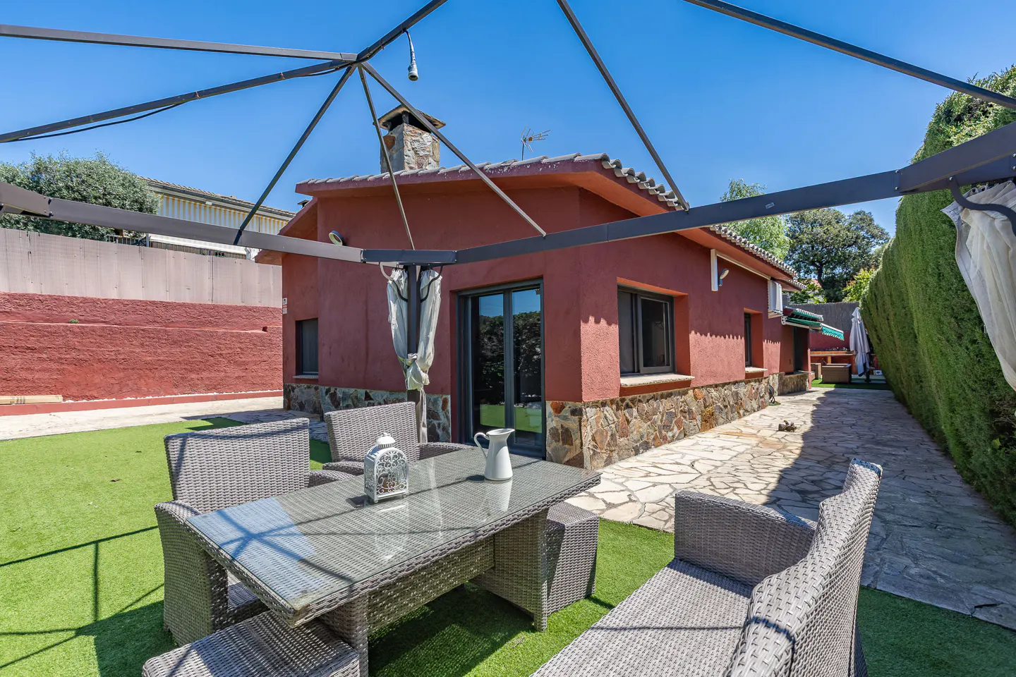 Outdoor patio with a glass table, wicker chairs, and a red house with a stone chimney in the background.