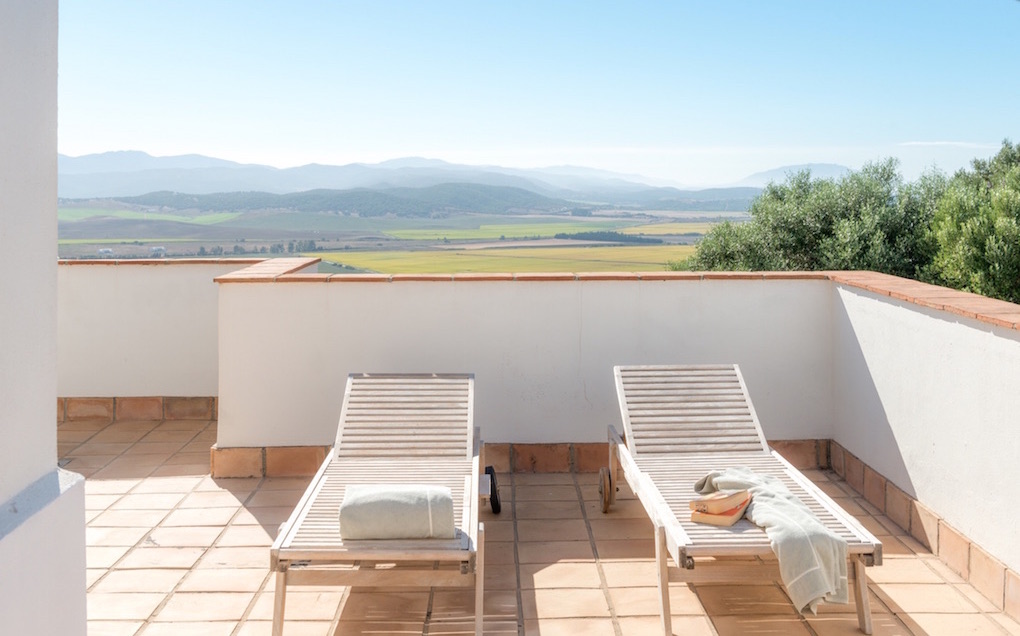 Two lounge chairs on a tiled patio overlook a green valley and distant mountains under a clear blue sky.