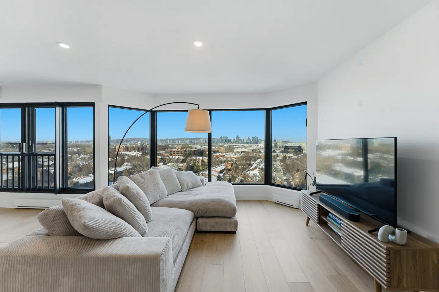 Bright living room with a large gray sectional sofa, floor lamp, and TV. Large windows offer a city view.