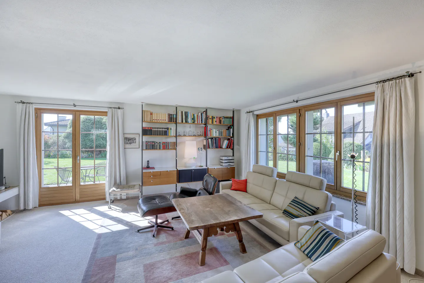 Bright living room with white sofa, wood table, and bookshelf. Large windows and doors lead to a green garden.