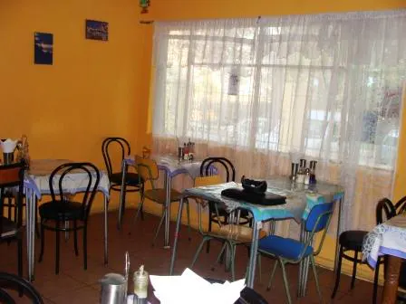 Interior view of a small restaurant with yellow walls, tables, and chairs. A large window with white curtains is visible.