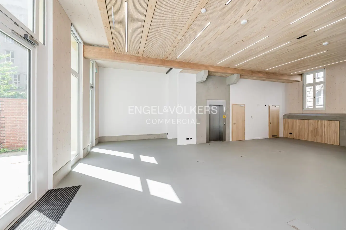Bright, empty commercial space with gray floor, wood ceiling, and large windows. An elevator and two doors are visible on the back wall.