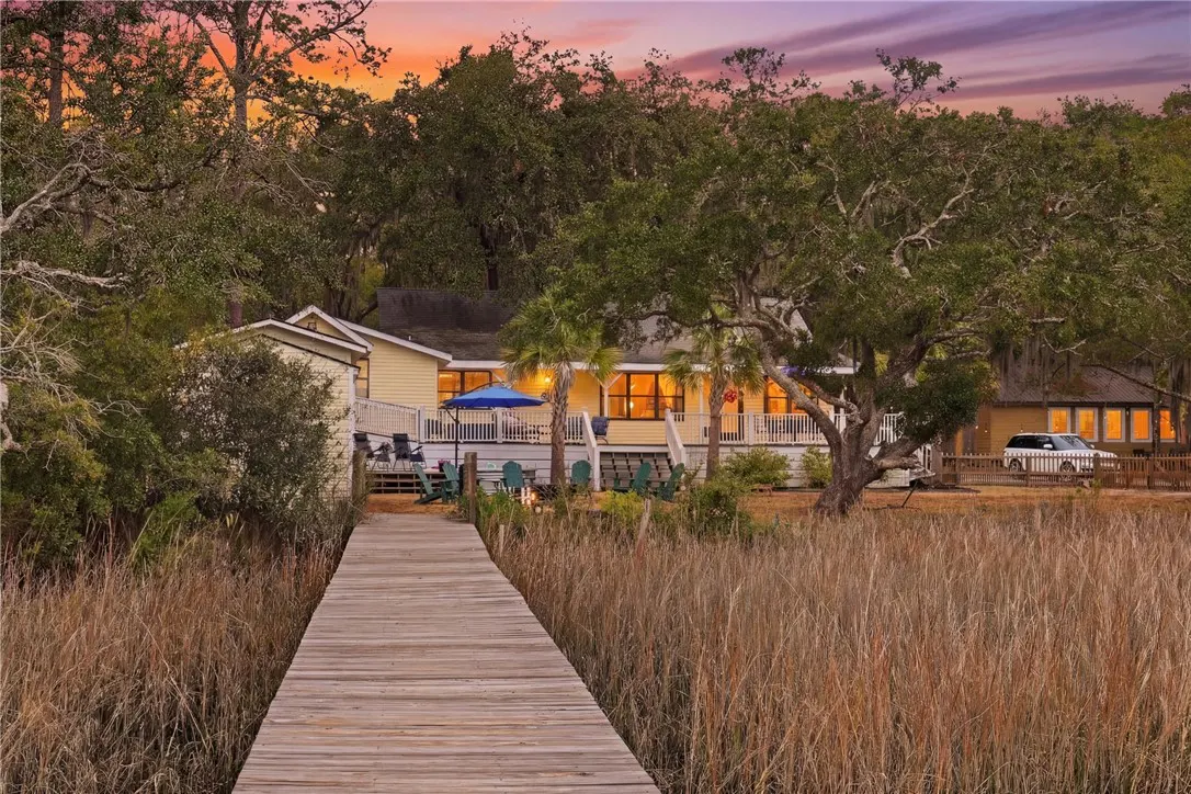 Sunset view of a yellow waterfront home with a wooden dock leading to it through tall grass. A deck with chairs and a blue umbrella are visible.