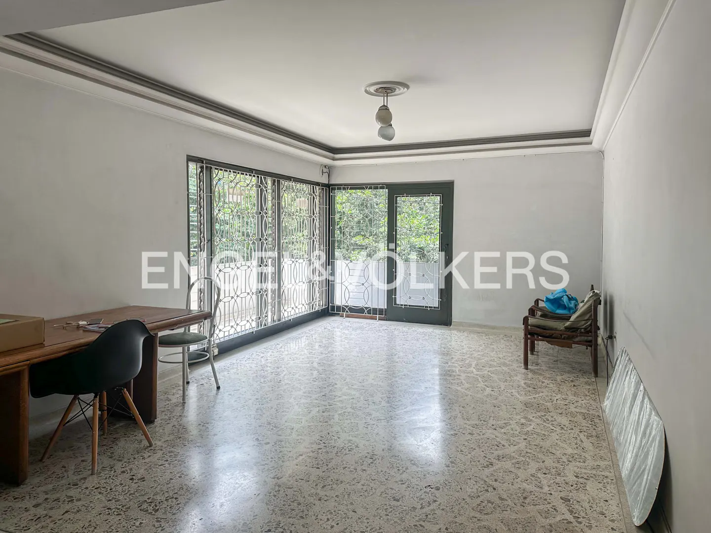 A bright room with a desk, chair, and bench. Large windows with metal grates look out to greenery. The floor is speckled tile.
