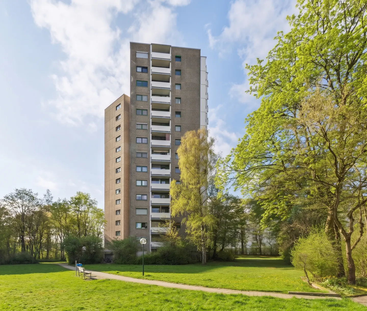 Exterior shot of a tall, gray apartment building with white balconies, surrounded by green trees and a grassy lawn.