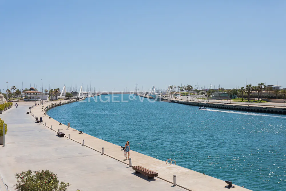 Scenic view of a marina with turquoise water, boats, and a concrete walkway with benches under a clear blue sky.