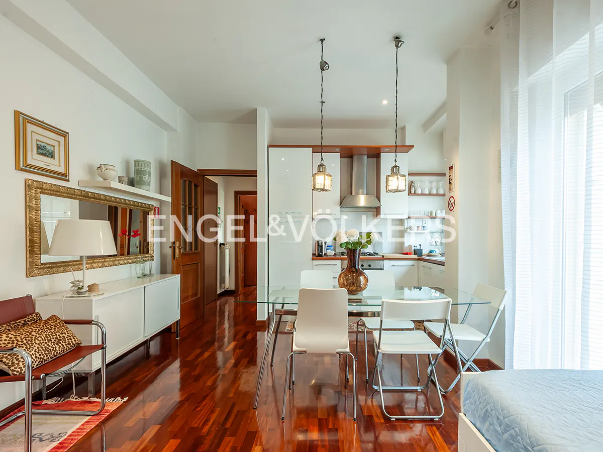 Bright apartment interior with wood floors, white walls, and a glass dining table with white chairs. Kitchen in the background.